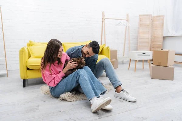 Happy interracial couple looking at bengal cat near carton boxes on floor in living room — стоковое фото