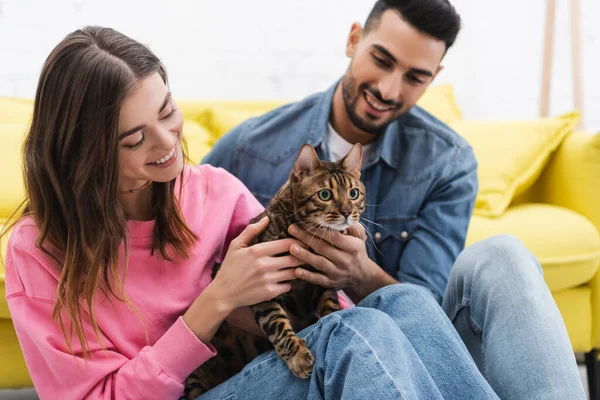 Cheerful woman petting bengal cat near blurred muslim man at home — Fotografia de Stock