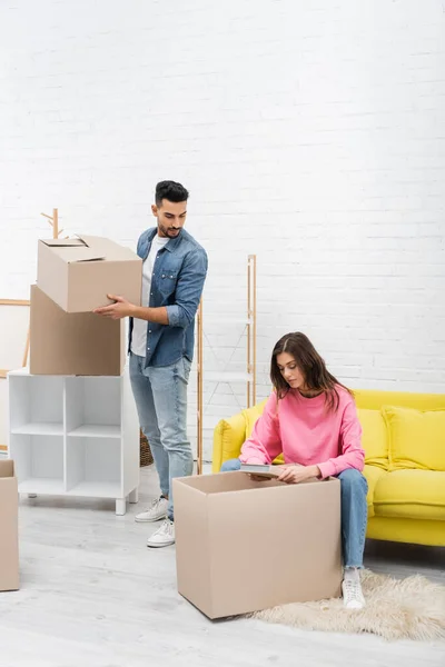 Woman holding book near muslim boyfriend with carton box at home — Stock Photo