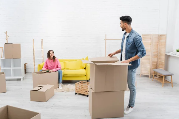 Cheerful woman holding book near arabian boyfriend and packages at home — Foto stock