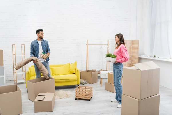 Fröhliche Frau hält Pflanzen neben muslimischen Freund mit Büchern und Teppich zu Hause — Stockfoto