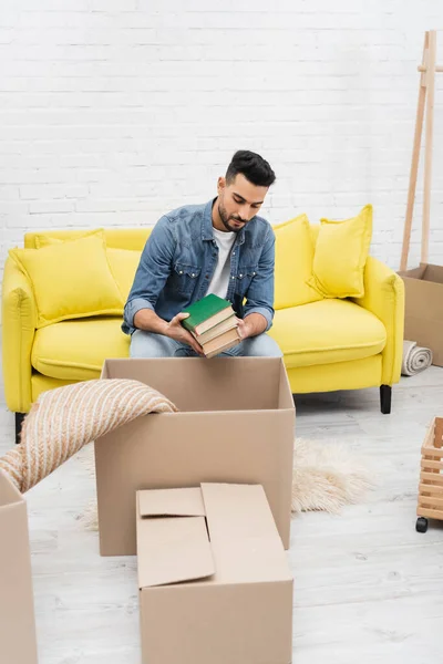 Young arabian man holding books near carton boxes and carpet at home — Stock Photo