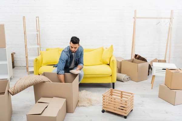 Arabian man unpacking package near boxes and bengal cat at home — Stock Photo