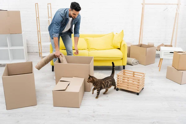 Arabe homme tenant tapis près des boîtes et chat bengale à la maison — Photo de stock