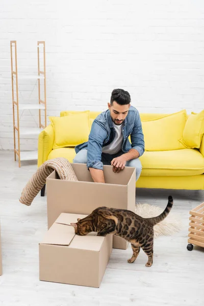 Muslim man unpacking cardboard box near bengal cat at home — стоковое фото