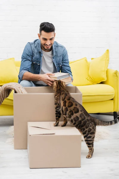 Cheerful arabain man holding books near bengal cat and packages at home — Stock Photo