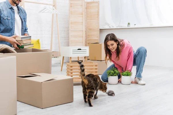 Mulher feliz olhando para bengala gato perto de plantas e namorado com pacotes em casa — Fotografia de Stock