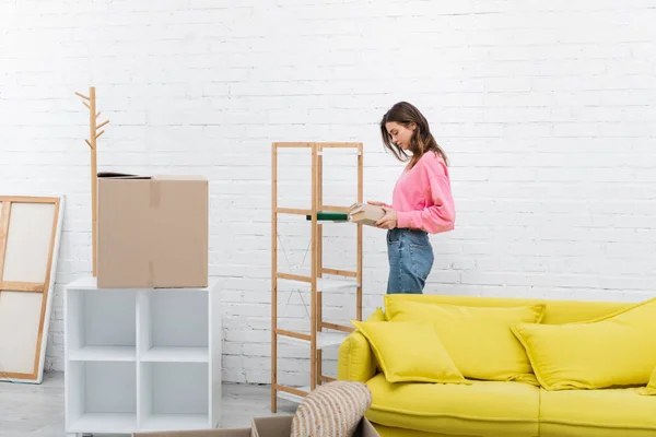 Side view of woman holding books near carton boxes and couch at home — стоковое фото