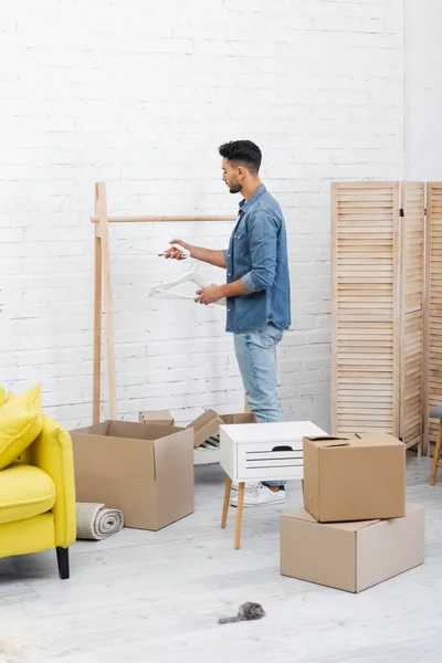 Vue latérale de l'homme musulman tenant des cintres près de rack et boîtes en carton à la maison — Photo de stock