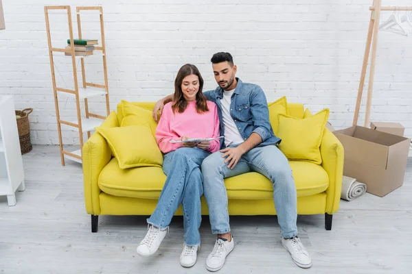 Sonriente pareja interracial sosteniendo paleta de colores en el sofá en casa - foto de stock