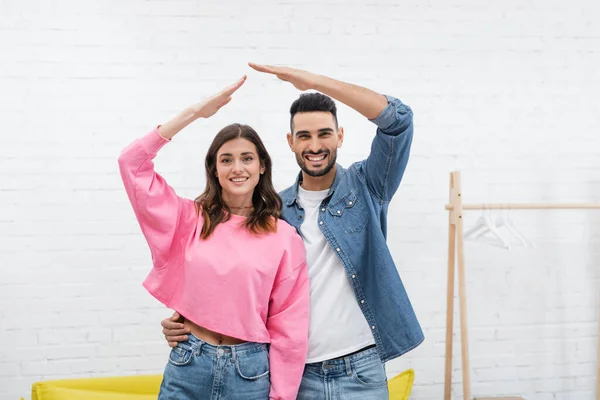 Positive multiethnic couple looking at camera and making roof gesture - foto de stock