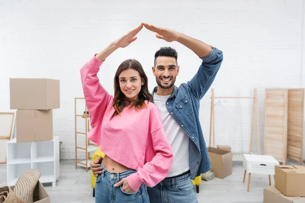 Positive interracial couple looking at camera near blurred carton boxes at home — Stock Photo