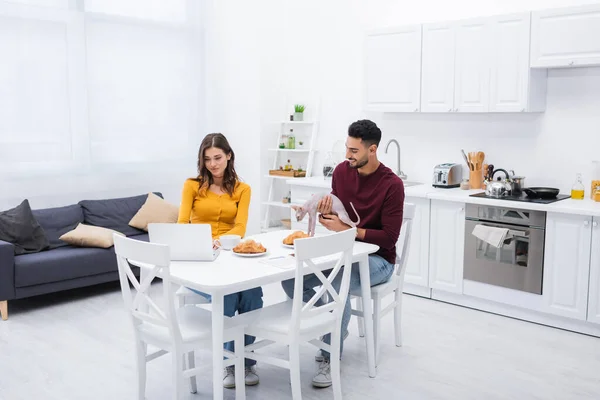 Smiling muslim man holding sphynx cat near breakfast and girlfriend with laptop at home — Foto stock