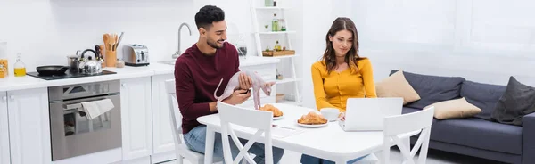 Muslim man holding sphynx cat near breakfast and girlfriend with laptop in kitchen, banner — Stock Photo