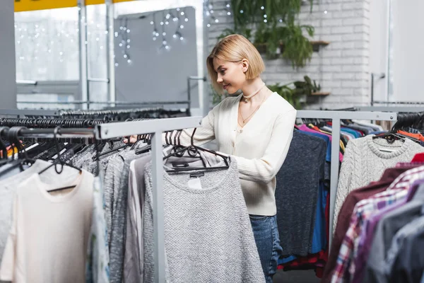 Side view of smiling woman looking at clothes on hangers in second hand — Stock Photo