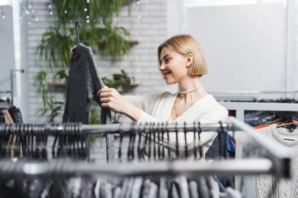 Seitenansicht einer fröhlichen Frau, die Pullover in der Nähe verschwommener Schienen in zweiter Hand hält — Stockfoto