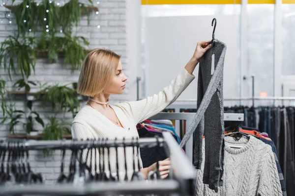 Вид сбоку на молодую женщину, держащую одежду возле вешалок в винтажном магазине — стоковое фото