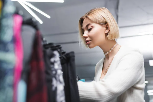 Young blonde woman choosing blurred clothes in second hand — стоковое фото