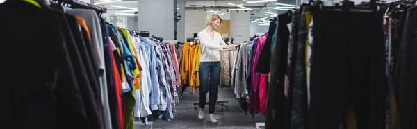 Vista lateral de la mujer positiva caminando cerca de la ropa en perchas en la tienda vintage, pancarta - foto de stock