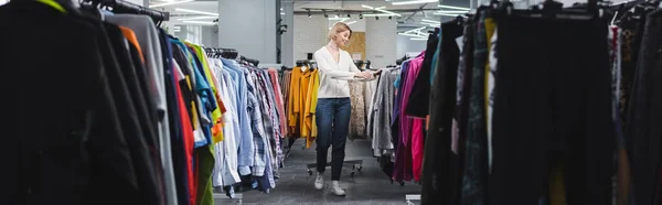 Vista laterale della donna positiva che cammina vicino ai vestiti su appendini in negozio vintage, banner — Foto stock