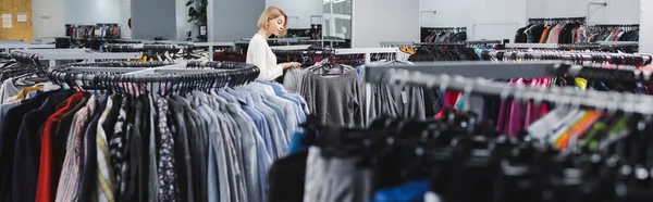 Side view of blonde woman choosing clothes on hangers in second hand, banner — стоковое фото