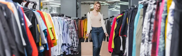 Mujer sonriente sosteniendo bolsas de compras cerca de la ropa en segunda mano, pancarta - foto de stock