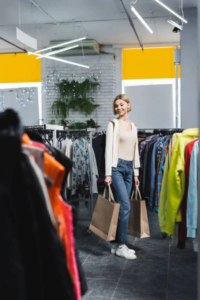 Cheerful woman holding purchases near clothes in second hand — Stock Photo