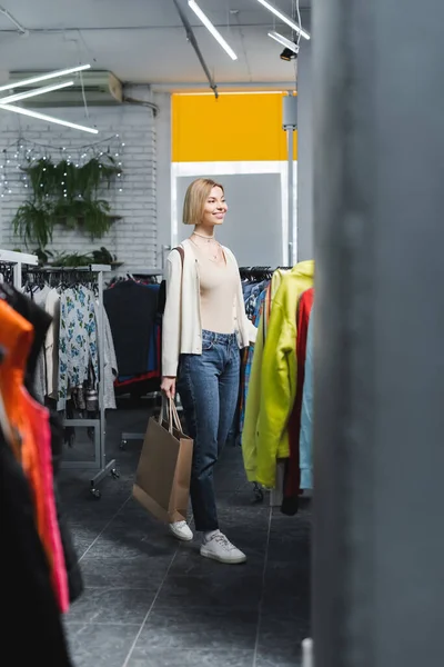 Mulher loira positiva segurando saco de compras perto de roupas em prateleiras em segunda mão — Fotografia de Stock
