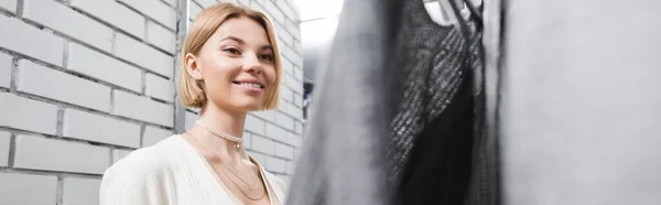 Positive woman looking at sweater in dressing room in vintage shop, banner — Stock Photo