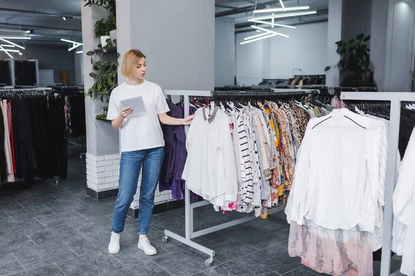 Retailer holding digital tablet near clothes in second hand — Stockfoto