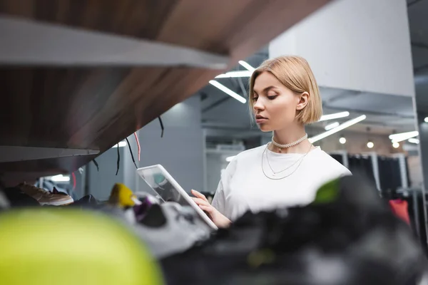 Blonde saleswoman using digital tablet near blurred shelf in second hand — Stock Photo