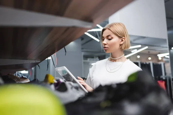 Blonde Verkäuferin benutzt digitales Tablet in der Nähe von verschwommenem Regal aus zweiter Hand — Stockfoto