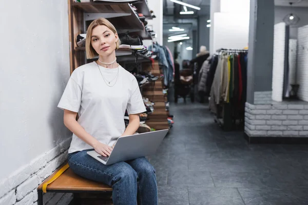 Blonde saleswoman holding laptop and looking at camera in vintage shop — Stockfoto