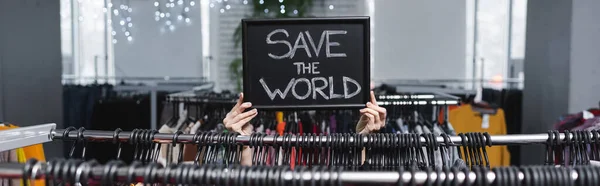 Ausgeschnittene Ansicht der Verkäuferin Haltebrett mit Rettet die Welt Schriftzug in der Nähe von Kleiderbügeln in Vintage-Shop, Banner — Stockfoto