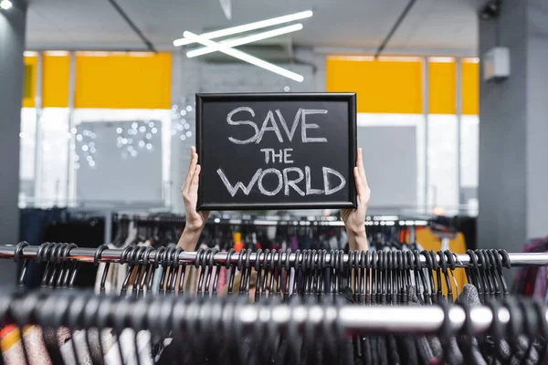 Cropped view of saleswoman holding nameplate with save the world lettering near clothes in second hand — стоковое фото