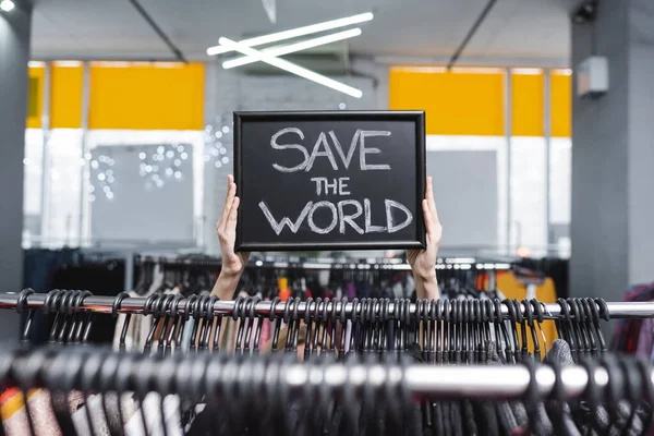 Cropped view of saleswoman holding nameplate with save the world lettering near clothes in second hand — Stock Photo
