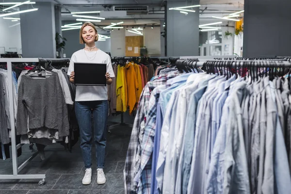 Smiling saleswoman holding empty board near clothes in second hand — Stockfoto