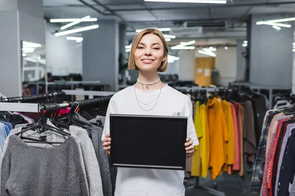 Vendedora positiva sosteniendo tablero vacío cerca de ropa borrosa en tienda vintage - foto de stock