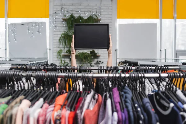 Verkäuferin hält leeres Brett in der Nähe von Kleiderbügeln in Vintage-Laden — Stockfoto