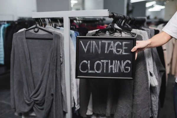 Vista ritagliata del rivenditore tenendo bordo con scritte di abbigliamento vintage vicino appendini di seconda mano — Foto stock