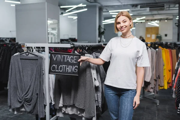 Lächelnde Verkäuferin hält Tafel mit Vintage-Klamotten-Schriftzug in Retro-Laden — Stockfoto