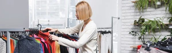 Side view of young customer choosing clothes near plants in second hand, banner — Stock Photo