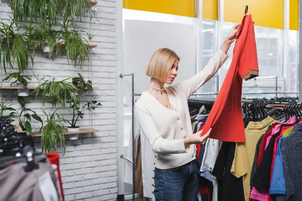 Bastante rubia cliente sosteniendo camiseta cerca de la ropa y las plantas durante las compras en la tienda vintage - foto de stock