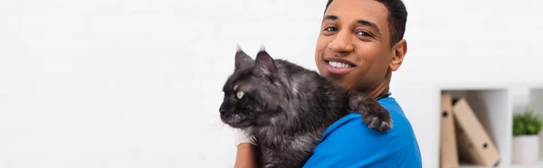 Fröhlich afrikanisch-amerikanischer Tierarzt hält Maine Coon in der Hand und blickt in die Kamera in der Klinik, Banner — Stockfoto