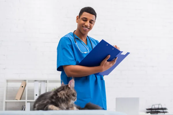 Lächelnder afrikanisch-amerikanischer Tierarzt schreibt auf Klemmbrett neben verschwommener Maine Coon Katze in Klinik — Stockfoto