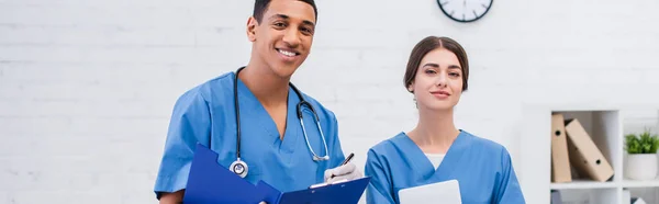 Cheerful interracial veterinarians with clipboard and digital tablet looking at camera in clinic, banner — Photo de stock