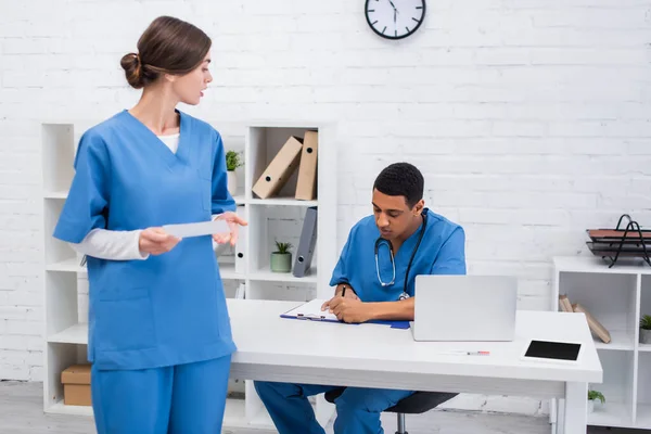 African american veterinarian writing on clipboard near colleague with scan in clinic — стокове фото