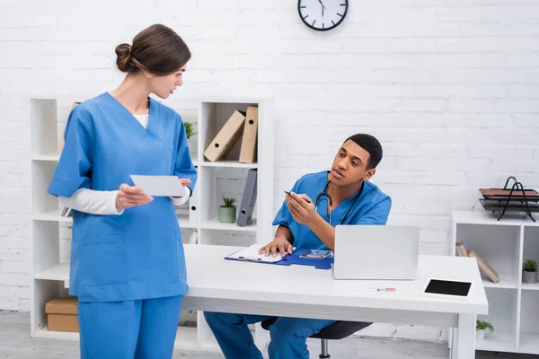 Médico veterinario afroamericano hablando con colega con exploración cerca del portapapeles y dispositivos en la clínica - foto de stock