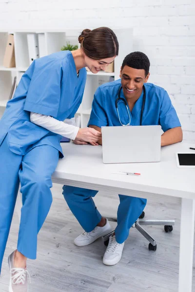 Médicos veteranos multiétnicos positivos mirando el portátil en la clínica - foto de stock