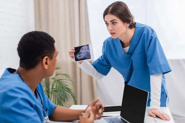 Vet doctor holding ultrasound scan of animal near blurred african american colleague in clinic - foto de stock